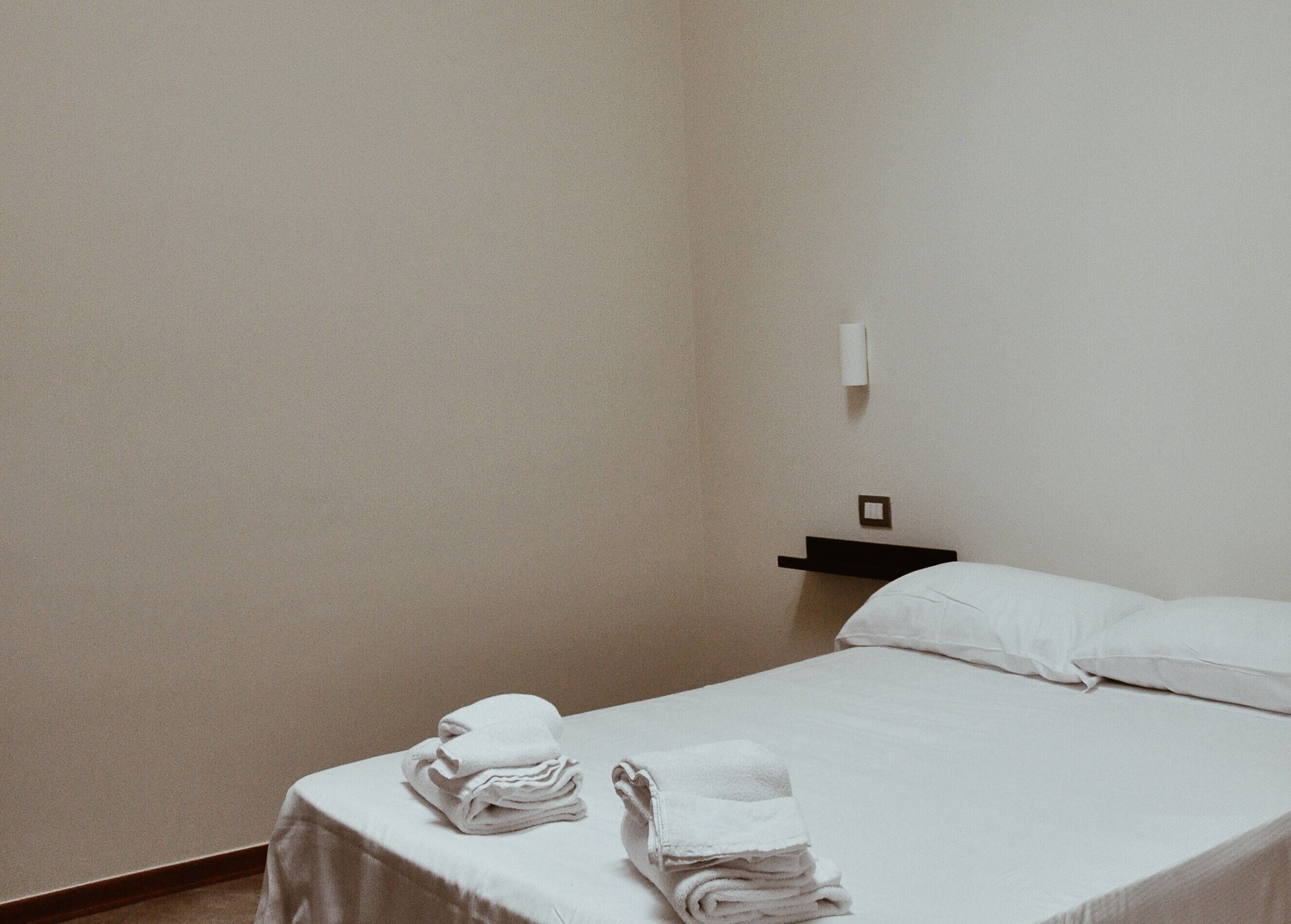 Une chambre d’hôtel simple et propre, avec un lit bien fait, une petite table de chevet et une lampe fixée au mur.