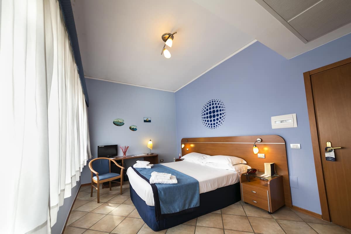 A hotel room with a bed, a desk with a computer, a chair, a bedside table, and a door.