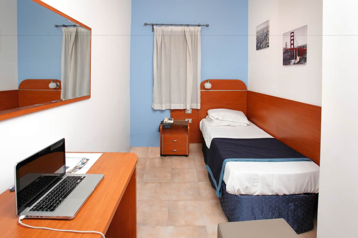A room with a bed, a desk with a laptop, a nightstand, and a mirror.