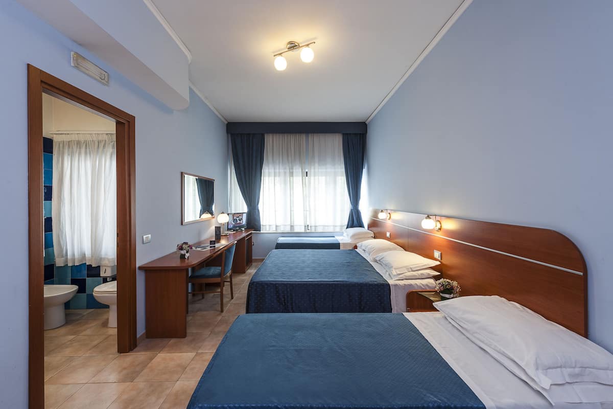A hotel room with two beds, a desk, a chair, a mirror, and a bathroom with a toilet and sink.