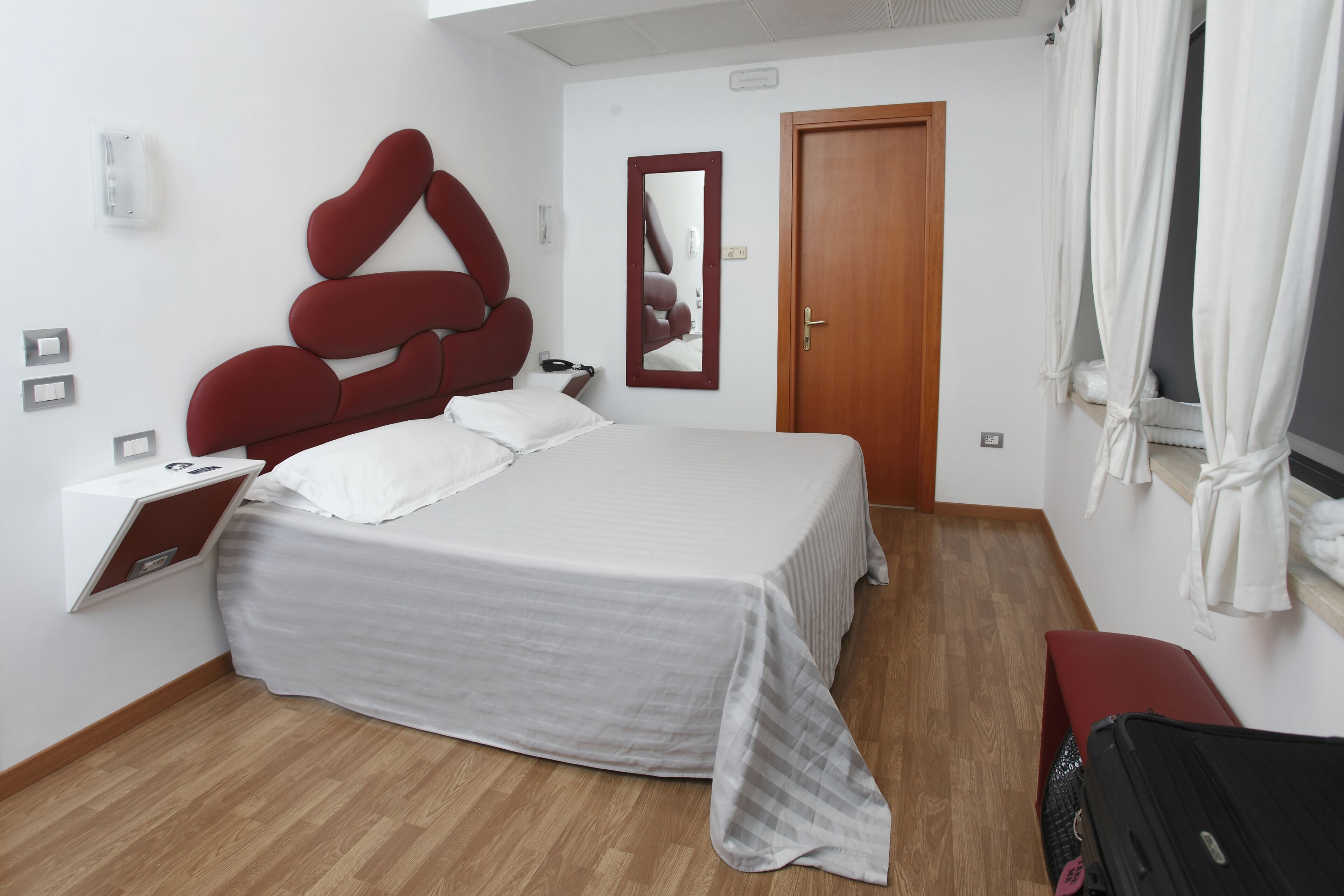 Ein Hotelzimmer mit einem Bett, Holzboden, einem roten Kopfteil, einem Spiegel, einer Tür und einem Fenster mit Vorhängen.