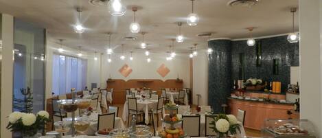 Uma sala de jantar com mesas redondas preparadas para uma refeição, iluminação pendente e uma área de cozinha ao fundo.