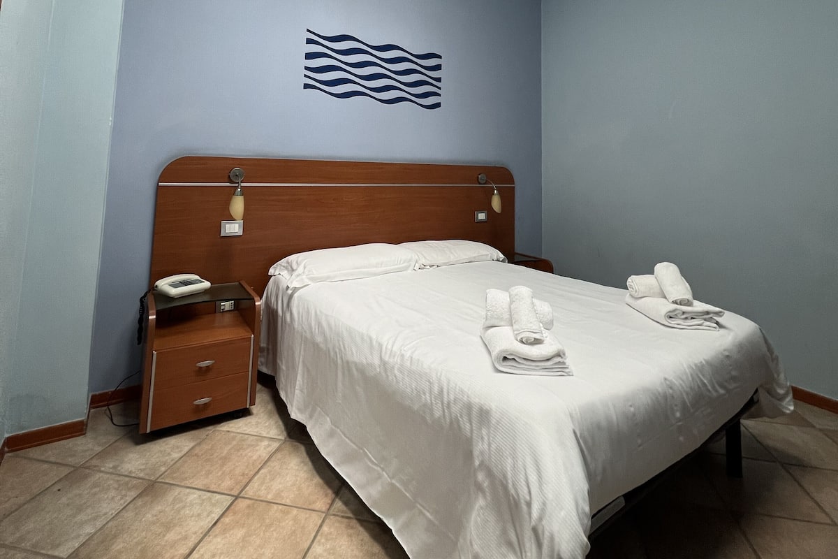 A hotel room with a wooden headboard, a bed with white linens, two rolled towels on the bed, a bedside table with a phone, and a wall-mounted light fixture.