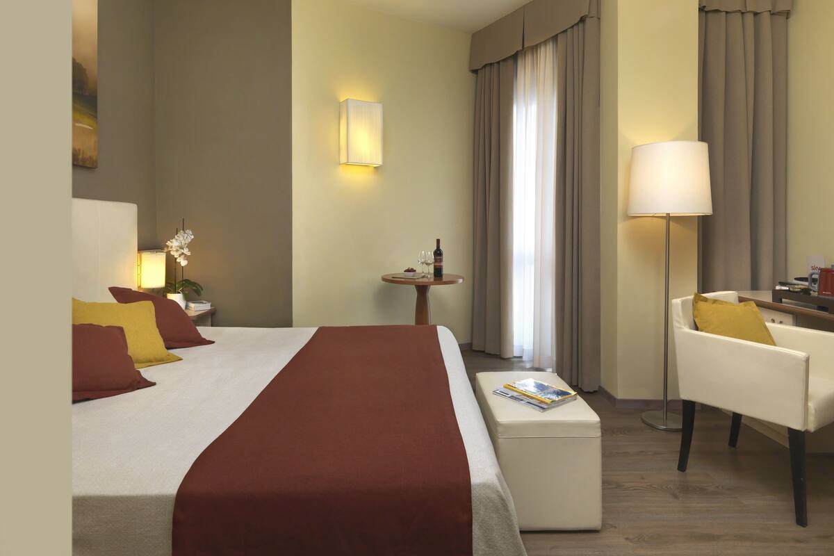 Une chambre d’hôtel avec un lit, une table de chevet avec une lampe, une petite table ronde avec une bouteille et des verres, une chaise avec un coussin et une grande fenêtre avec des rideaux.