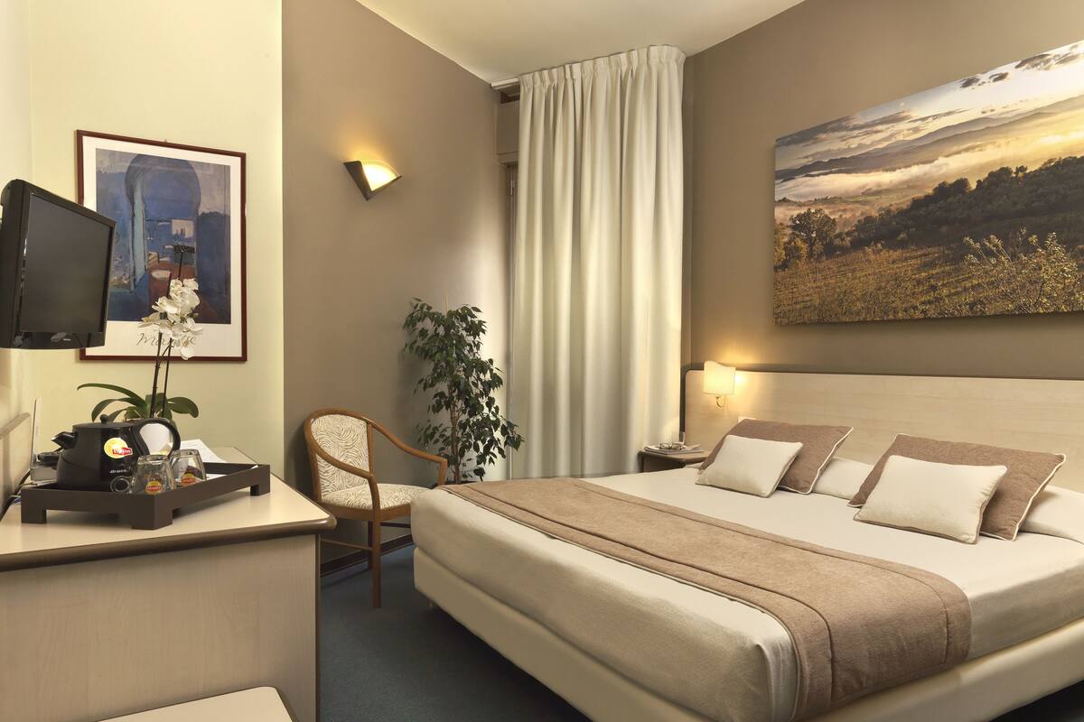 Une chambre d’hôtel avec un lit, un bureau doté d’une bouilloire, une chaise, une plante et un tableau au mur.