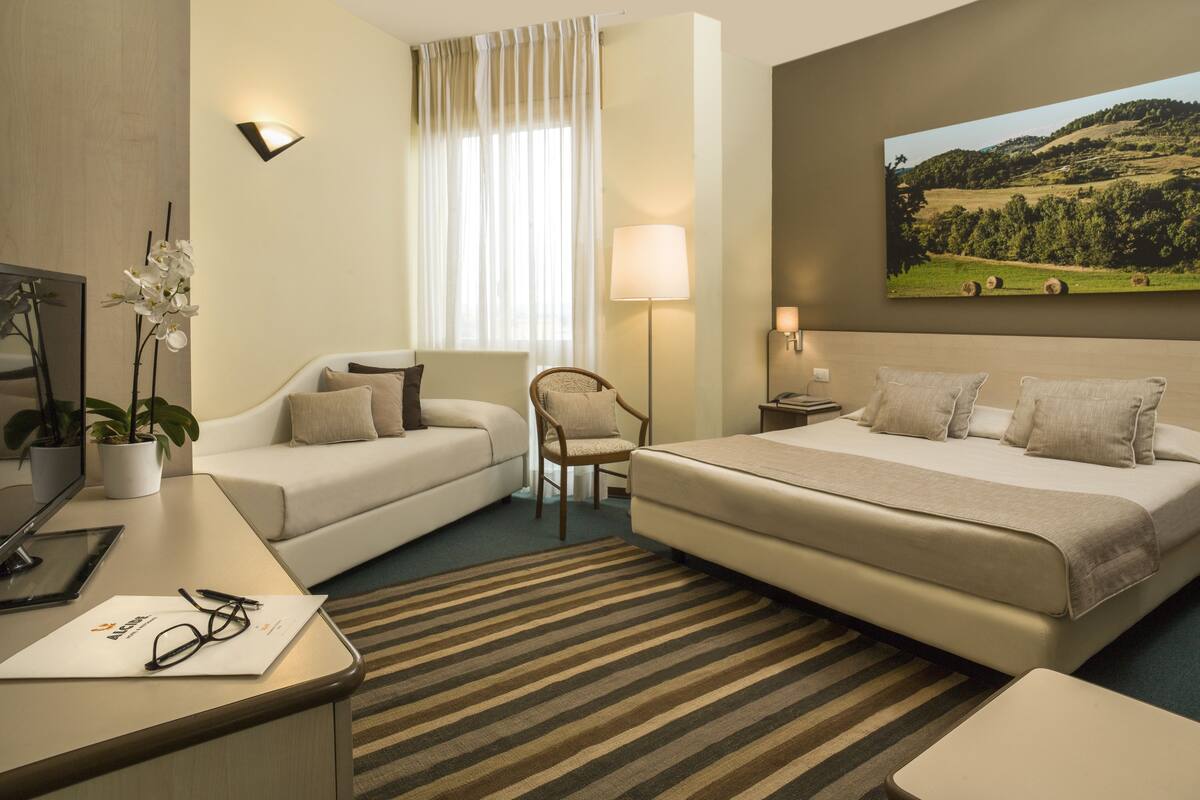 Une chambre d’hôtel avec un lit, un canapé, une chaise, un bureau avec une lampe, une plante en pot et un tableau au mur.