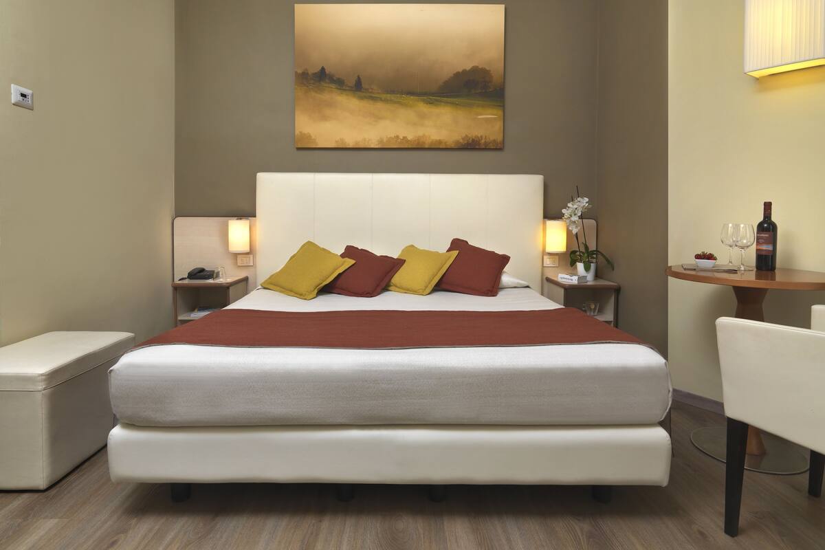 Une chambre d’hôtel avec un grand lit, une table de chevet avec une lampe, une petite table avec une bouteille et des verres, et un tableau au mur.