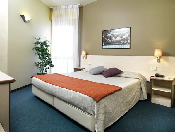 A hotel room with a bed, a nightstand, a lamp, a plant, and a painting on the wall.
