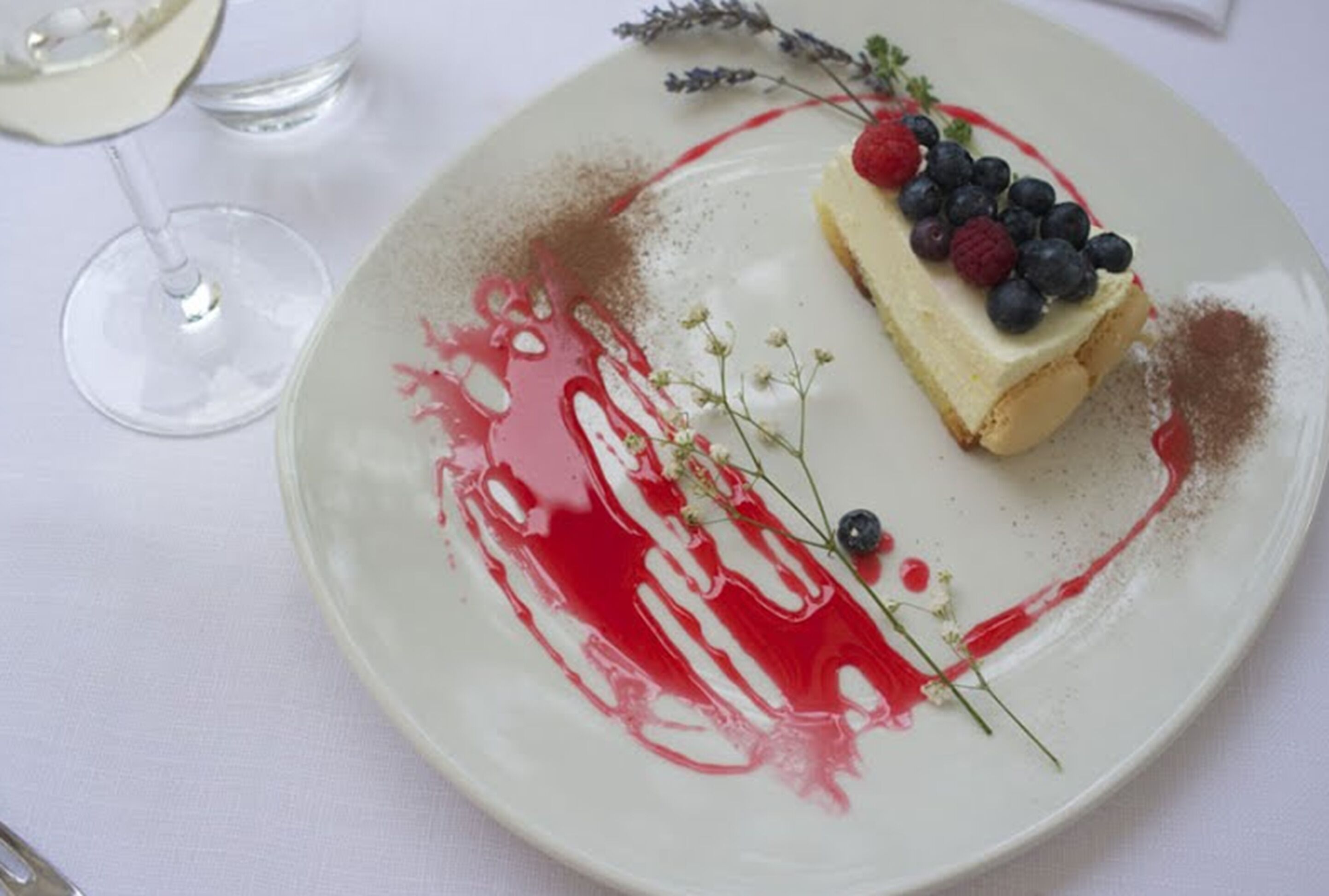 A dessert plate with a slice of cheesecake topped with berries and a drizzle of red sauce, accompanied by a glass of white wine.