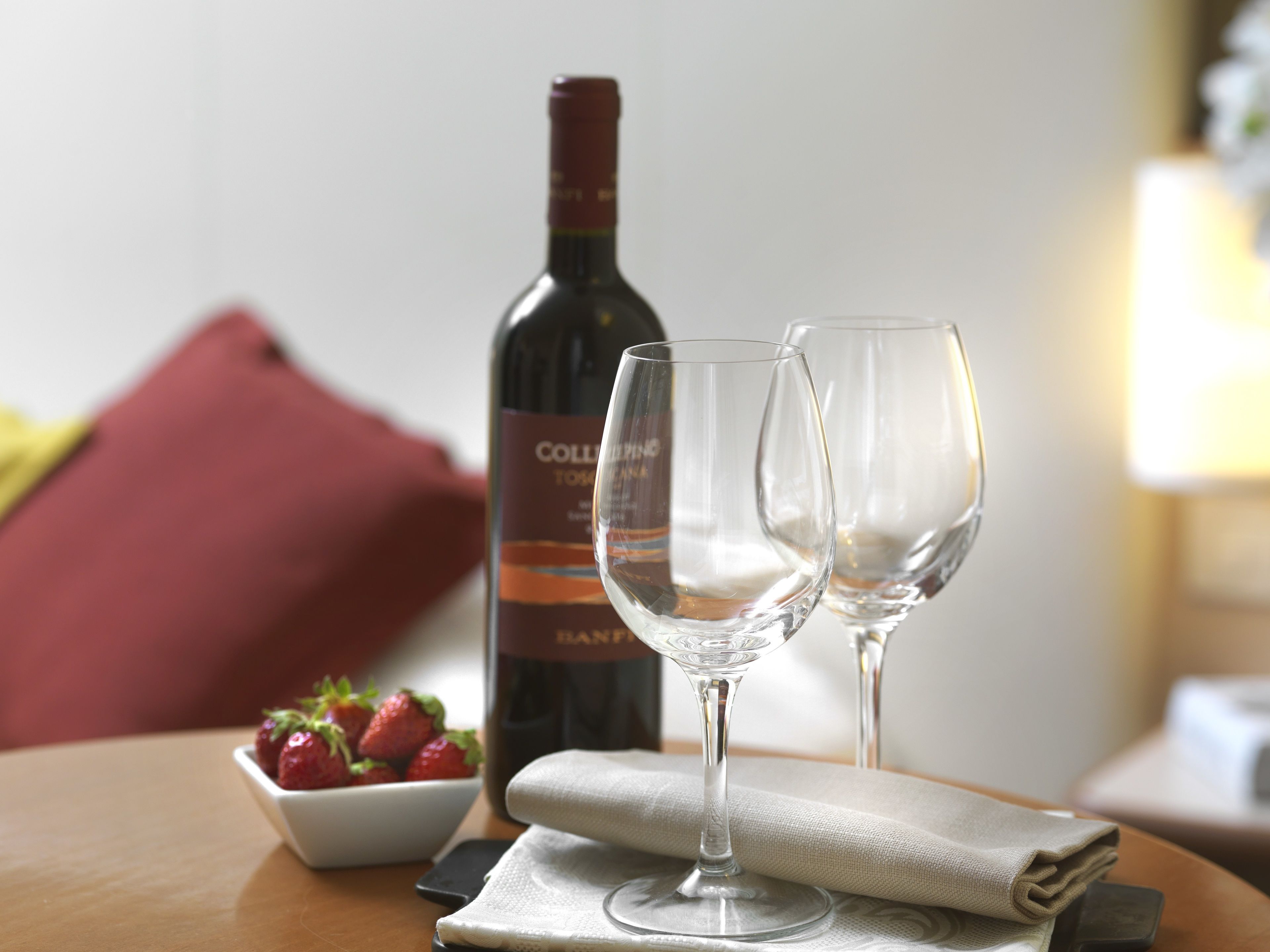 A bottle of wine, two empty wine glasses, a bowl of strawberries, and a smartphone on a wooden table.