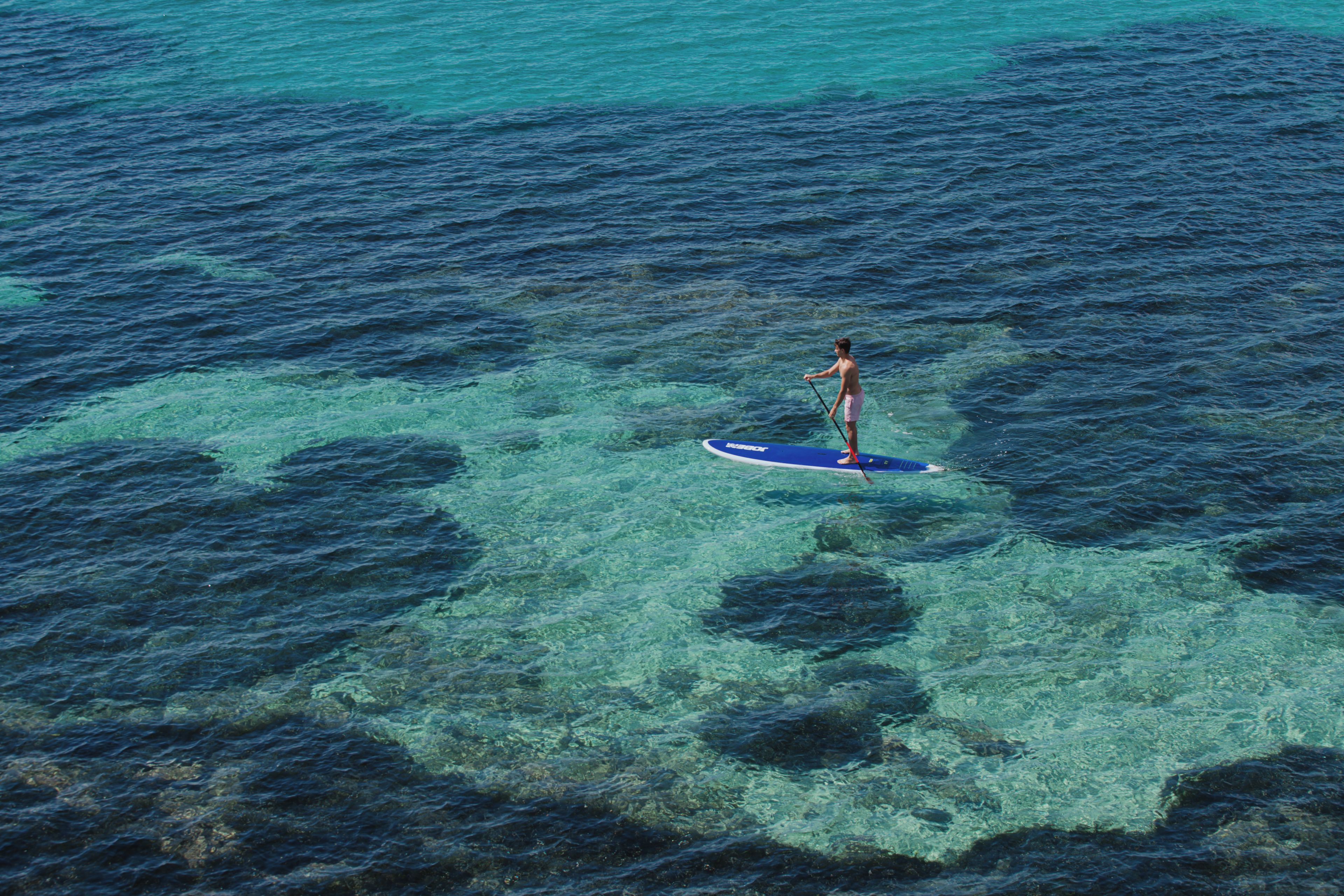 A person standing on a paddleboard in clear water with visible underwater rocks.