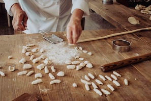 Iemand bereidt pasta aan een houten tafel, met een rasp, deegroller en metalen beker.