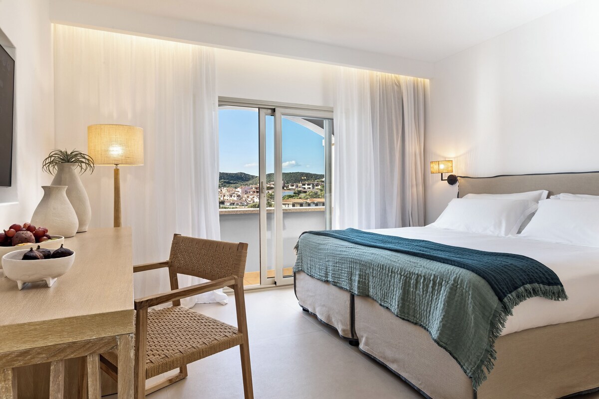 Een hotelkamer met een bed, een bureau met stoel, een lamp en uitzicht op een stadsgezicht door een glazen deur.