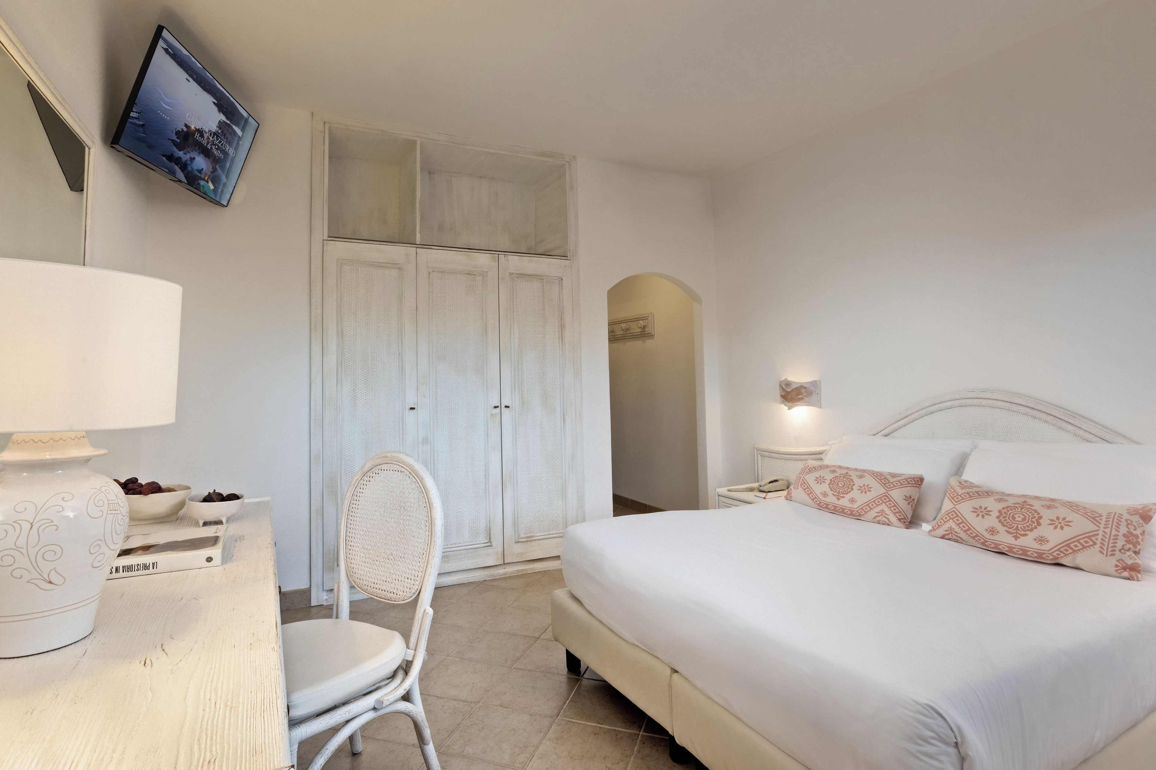 A hotel room with a bed, a desk with a lamp, a chair, a wardrobe, and a television on the wall.