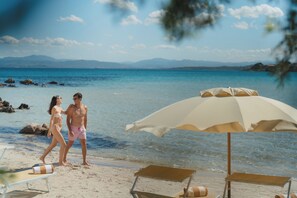 Une scène de plage avec un ciel dégagé, une mer calme et un couple qui se promène. On peut voir des chaises de plage et un parasol.