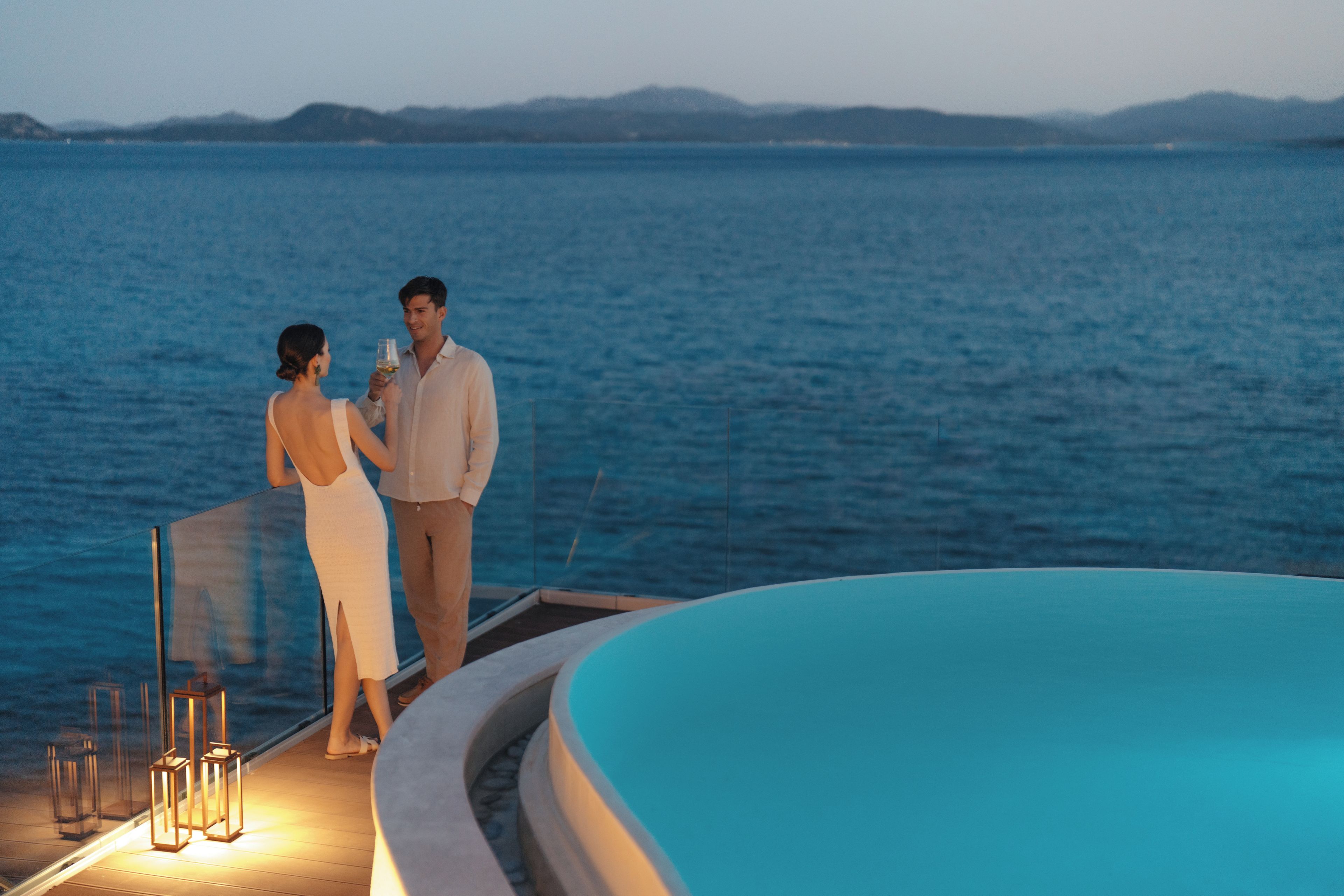 A couple standing by a pool with a view of the sea and distant mountains.