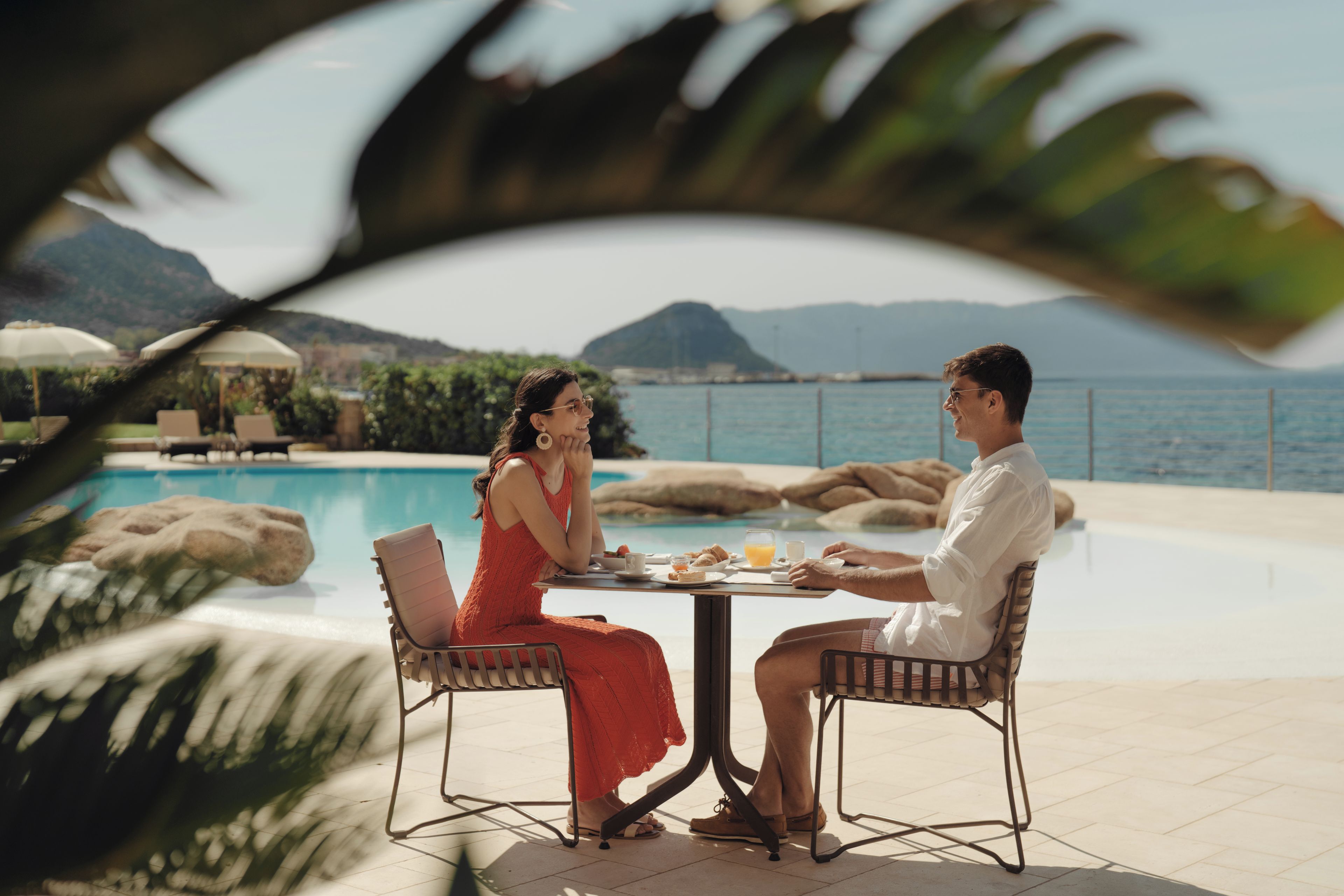 A couple dining at a table by a pool with a view of the sea and mountains.