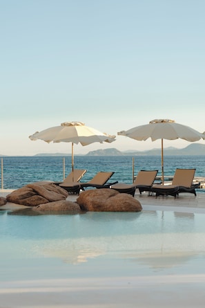 Un espace aménagé au bord de la piscine, avec des chaises longues et des parasols, offrant une vue sur une étendue d’eau et sur des montagnes lointaines.