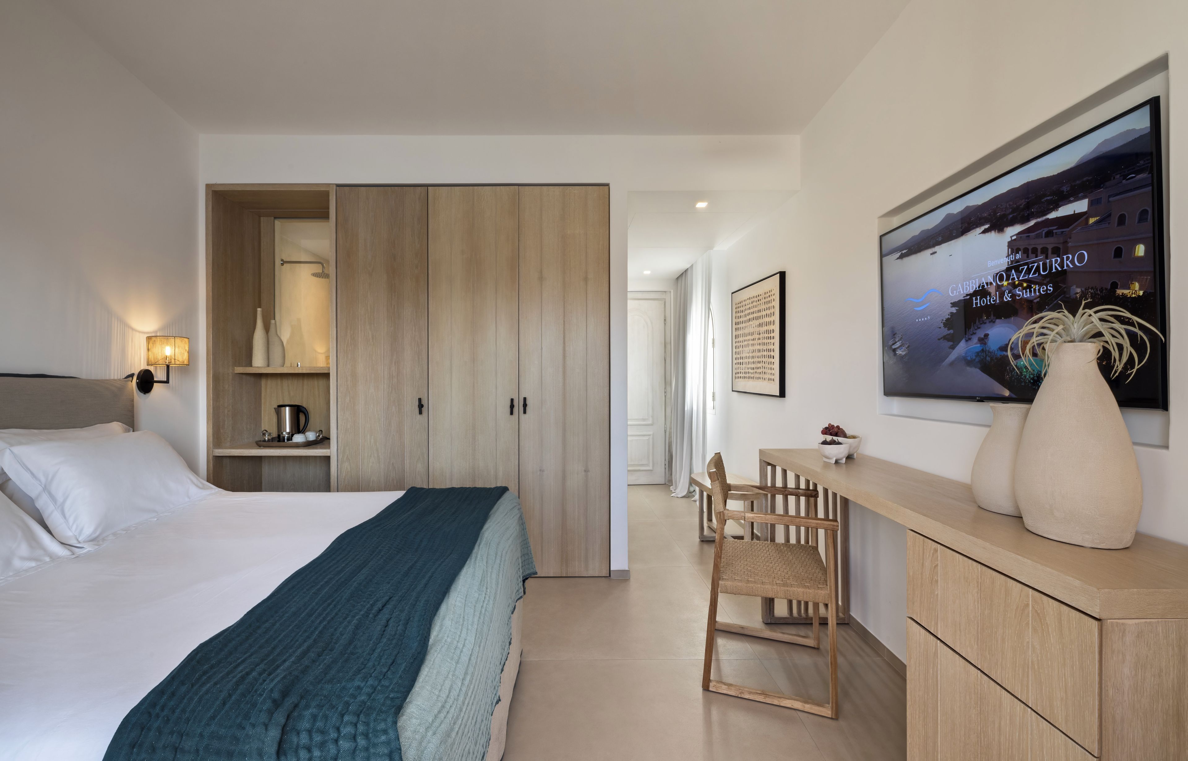 Een hotelkamer met een bed, een houten garderobe, een bureau met stoel, en een groot raam met uitzicht op een wateroppervlak.