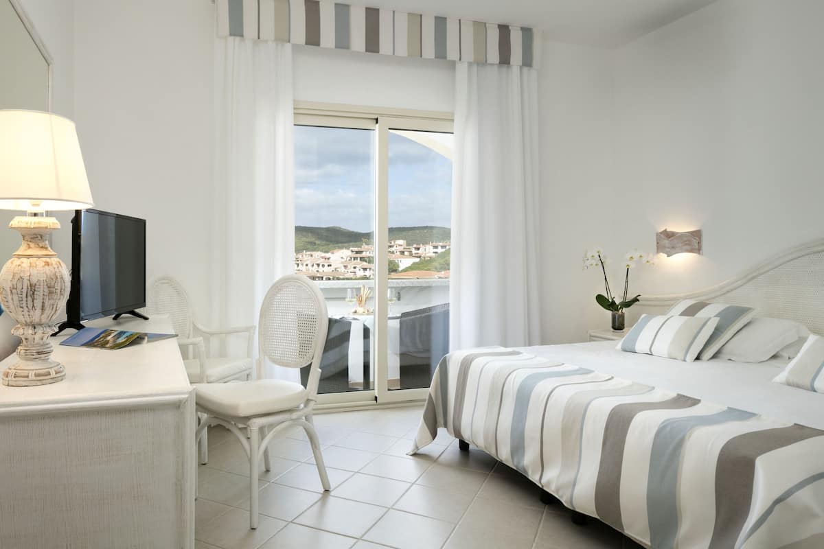Ein Hotelzimmer mit Bett, Schreibtisch mit Lampe, Sessel, Fernseher und Blick auf die Stadt durch das Fenster.