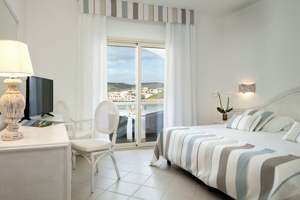 Une chambre d’hôtel avec un lit, un bureau doté d’une lampe, une chaise, un téléviseur et une vue sur la ville par la fenêtre.