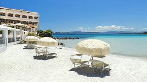 Une plage avec des parasols blancs et des chaises longues, un immeuble doté de balcons, et une vue dégagée sur la mer et les montagnes.