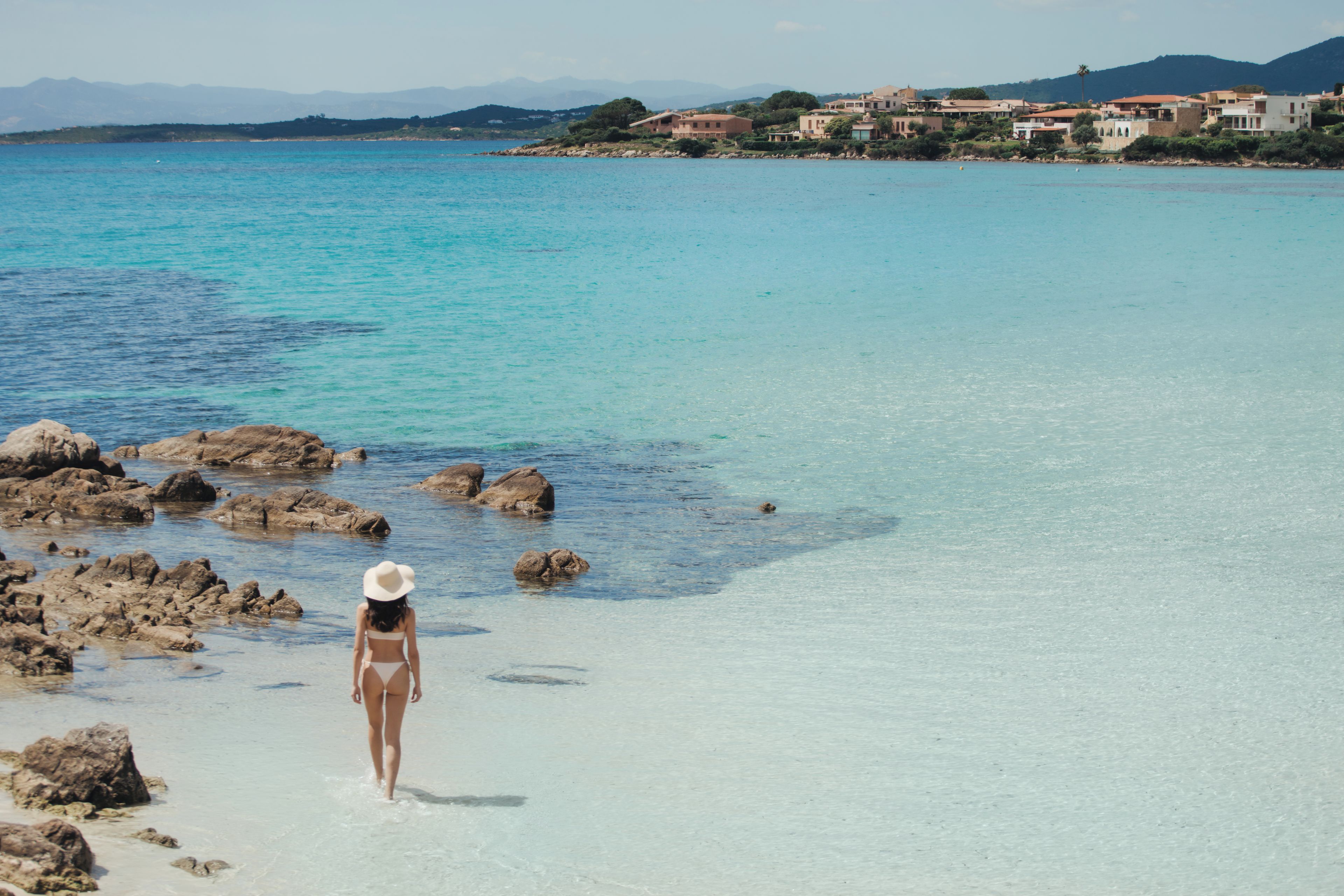 Eine Frau in einem Bikini geht über einen felsigen Strand mit klarem Wasser und einer Stadt im Hintergrund.