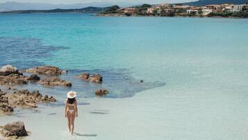 Eine Frau in einem Bikini geht über einen felsigen Strand mit klarem Wasser und einer Stadt im Hintergrund.