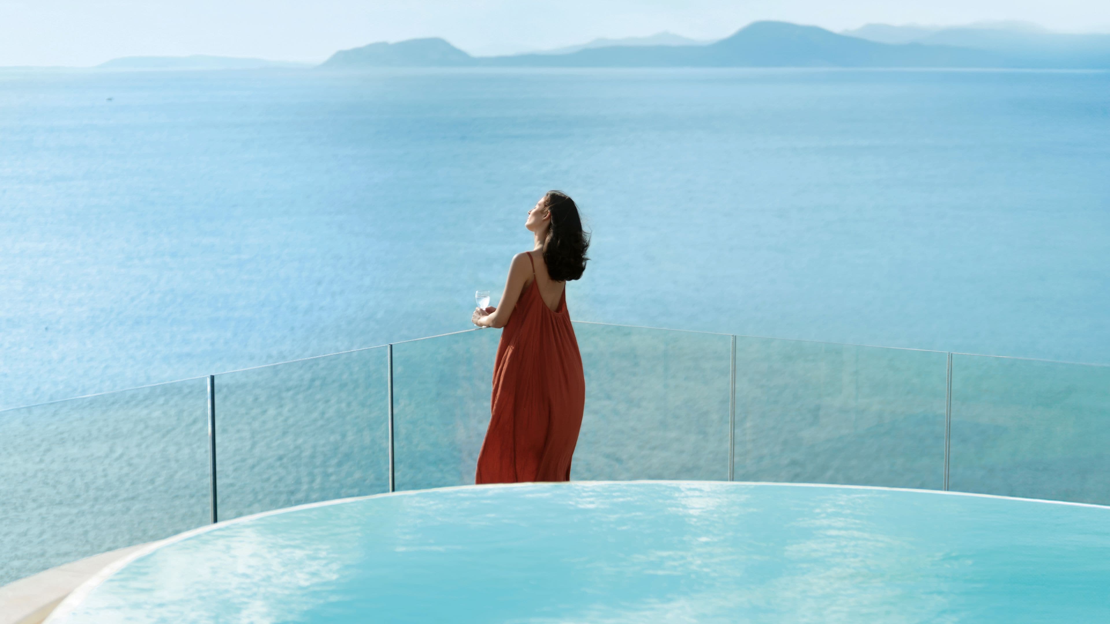 A person standing by a pool with a view of the sea and distant mountains.