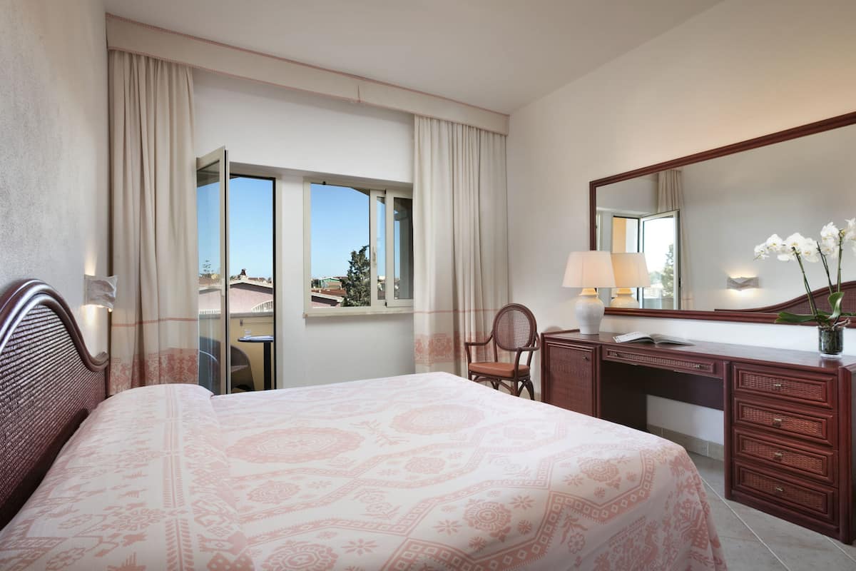 Ein Hotelzimmer mit einem großen Bett, einem Schreibtisch inklusive Spiegel, einem Sessel und einem Fenster mit Vorhängen.