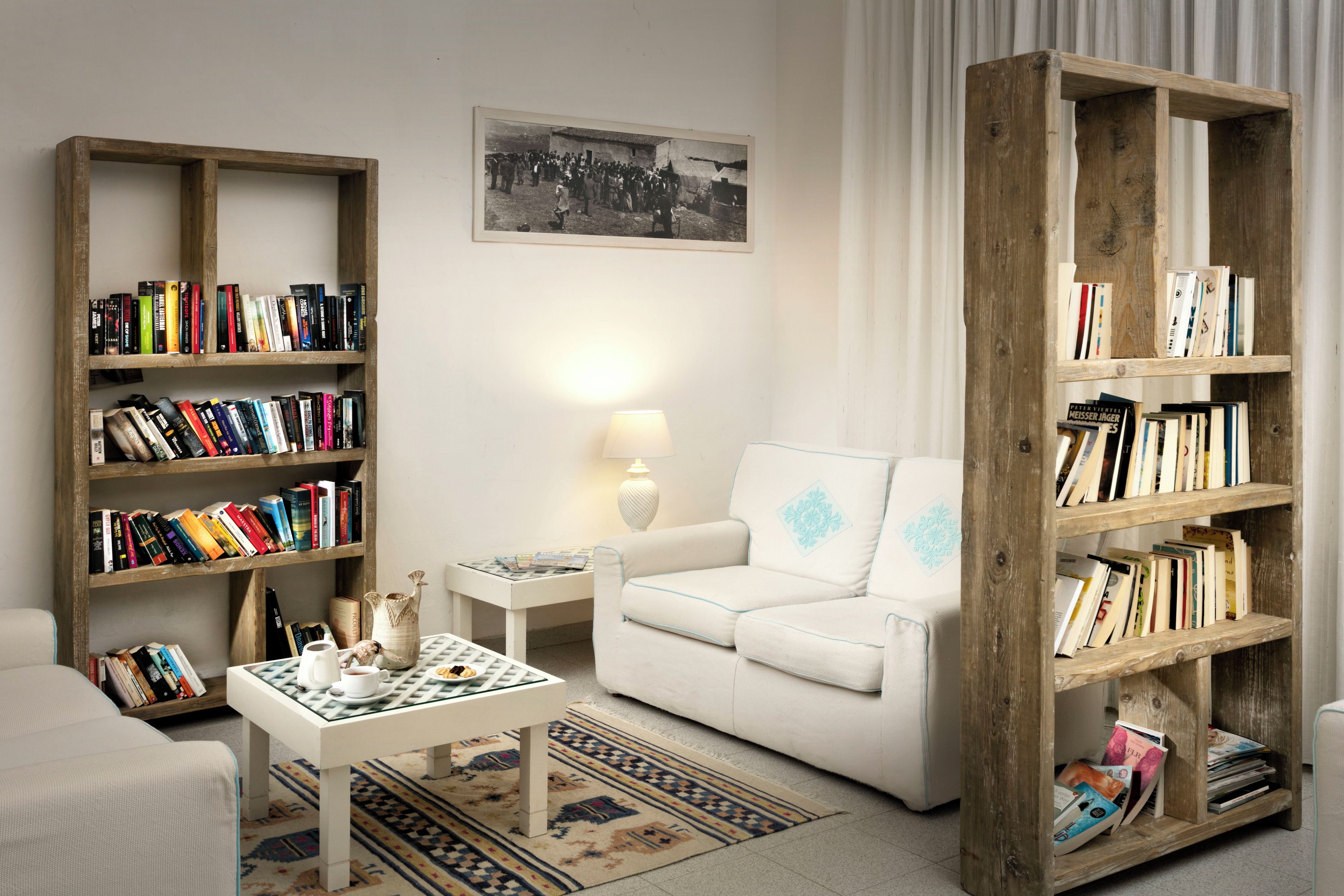 A living room with a white sofa, two wooden bookshelves filled with books, a small table with a lamp, and a patterned rug.