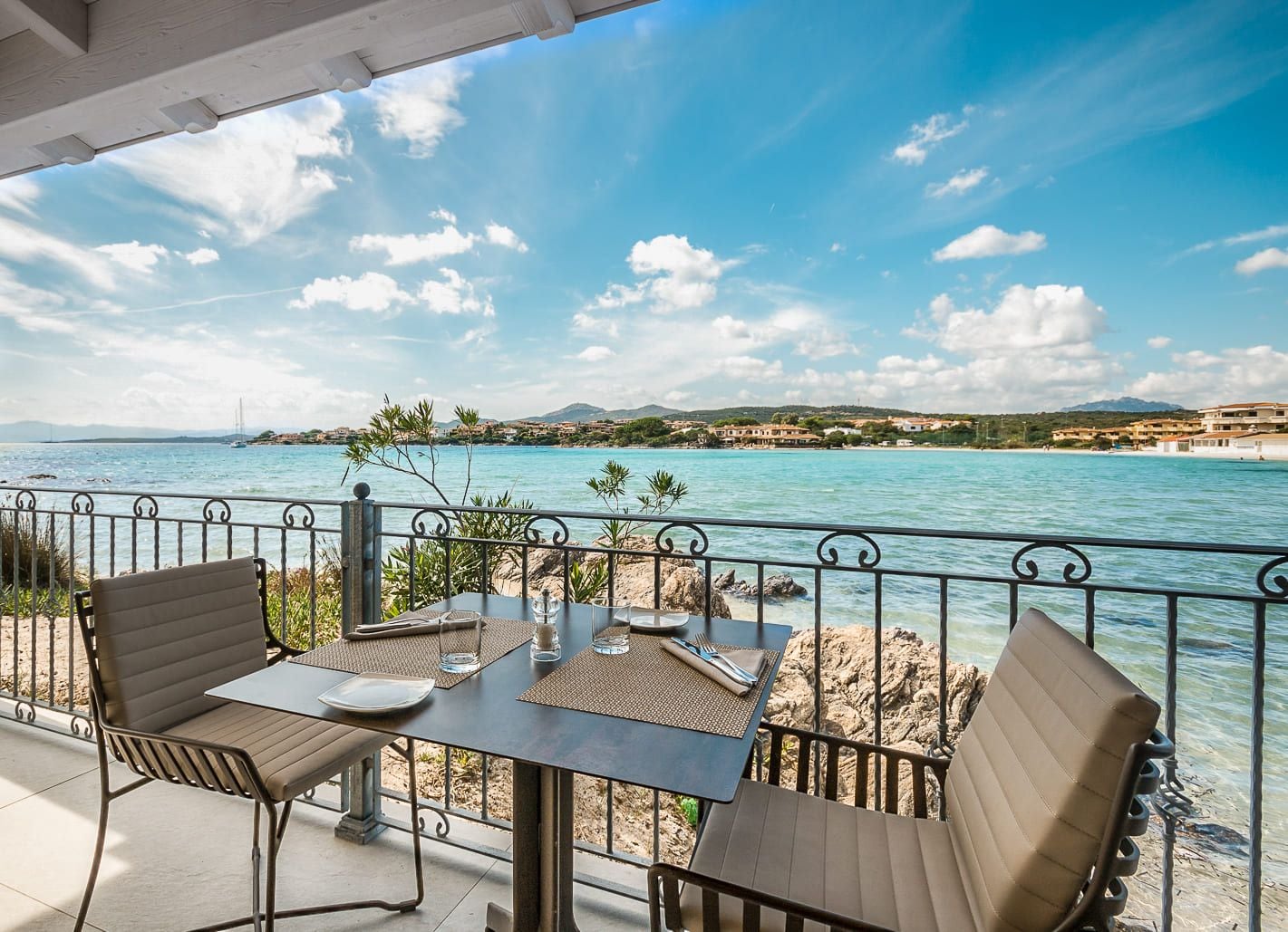 A seaside terrace with a table and chairs, overlooking a calm sea and a coastal town.