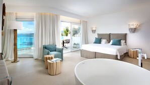 Een moderne hotelkamer met een groot bed, een zithoek met een stoel en bijzettafels, een badkuip en een balkon met uitzicht op zee.