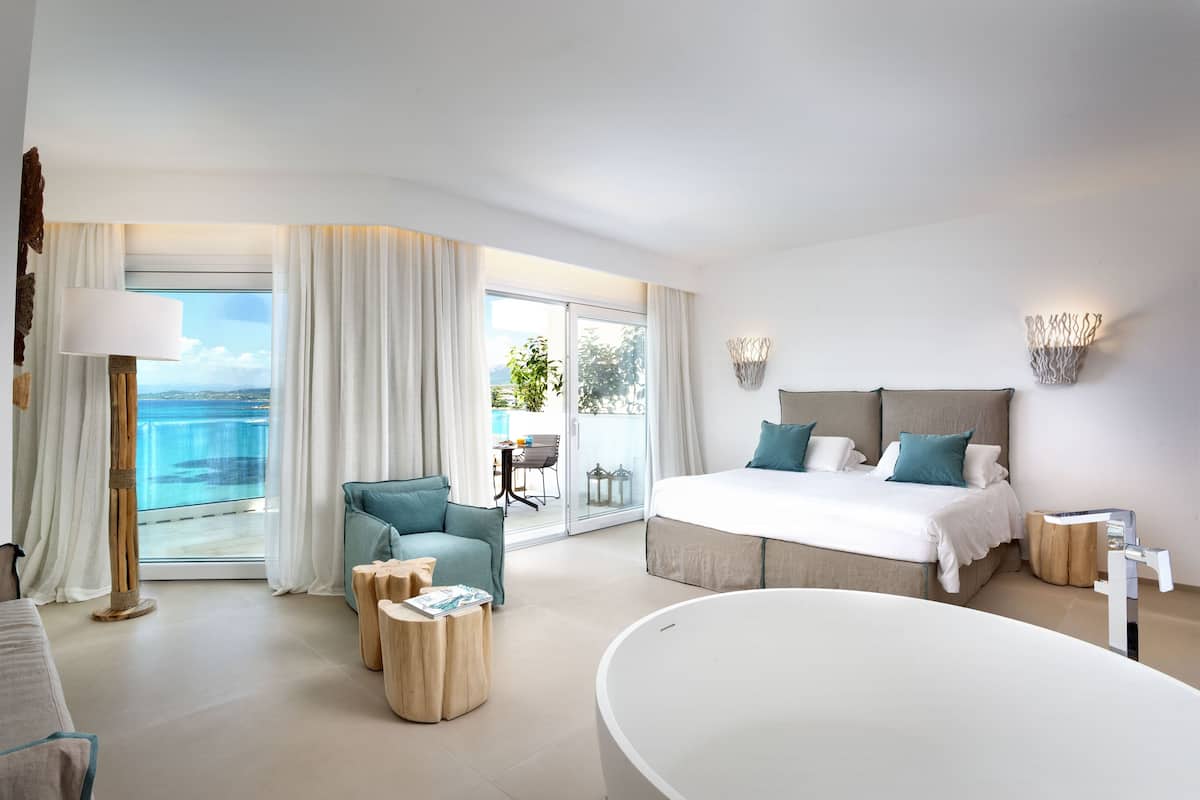Ein modernes Hotelzimmer mit einem großen Bett, einer Sitzecke mit Sessel und Beistelltischen, einer Badewanne und einem Balkon mit Meerblick.