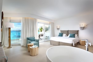 Una habitación de hotel moderna con una cama grande, un área de estar con una silla y mesitas auxiliares, una bañera y un balcón con vista al mar.
