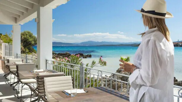 Une femme, vêtue d’une chemise blanche et d’un chapeau, se tient sur un balcon d’où l’on aperçoit la mer et les montagnes.