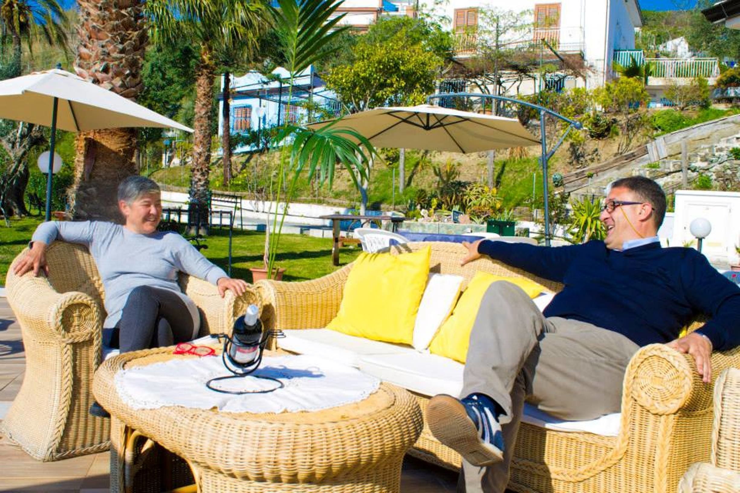Un fauteuil de jardin en osier, accompagné d’une table blanche et de coussins jaunes, installé à l’extérieur, sous des parasols et à proximité de palmiers.