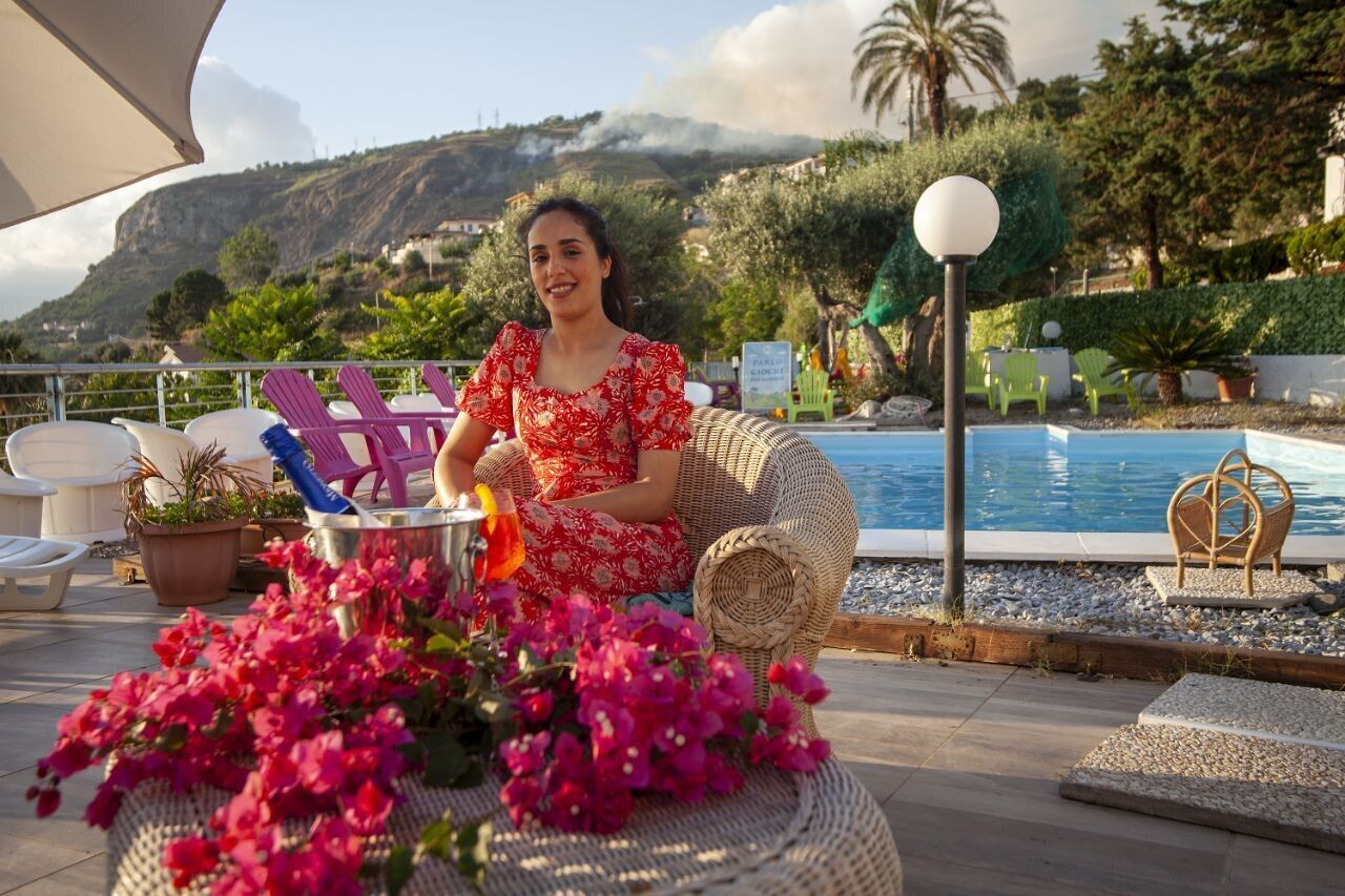 A woman sitting on a wicker chair by a pool with a drink, surrounded by potted plants and a view of a mountainous landscape.