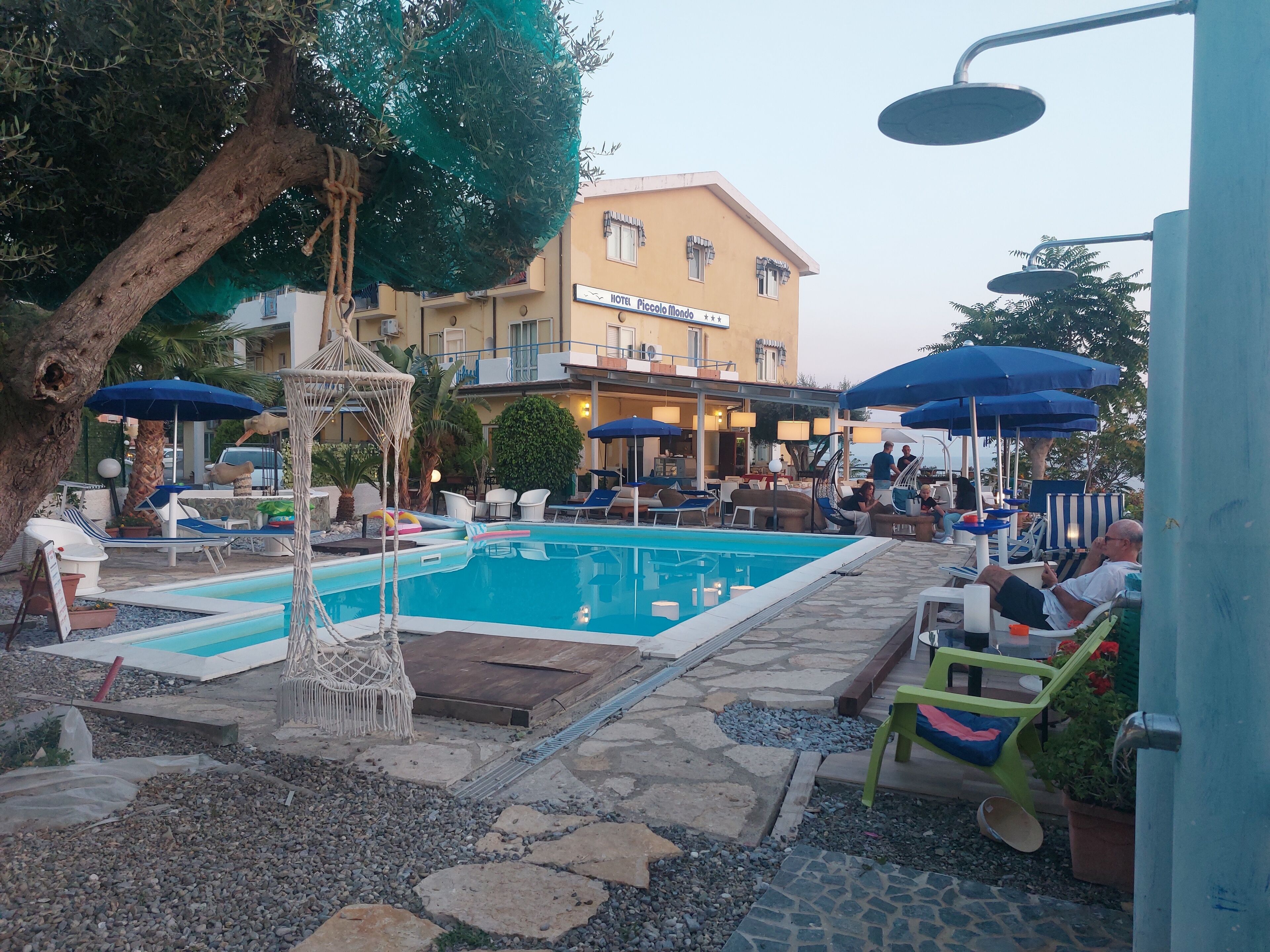 A pool area with lounge chairs and umbrellas, a building with a sign reading "HOTEL PICCOLO FILANDO", and a tree with a hanging decoration.