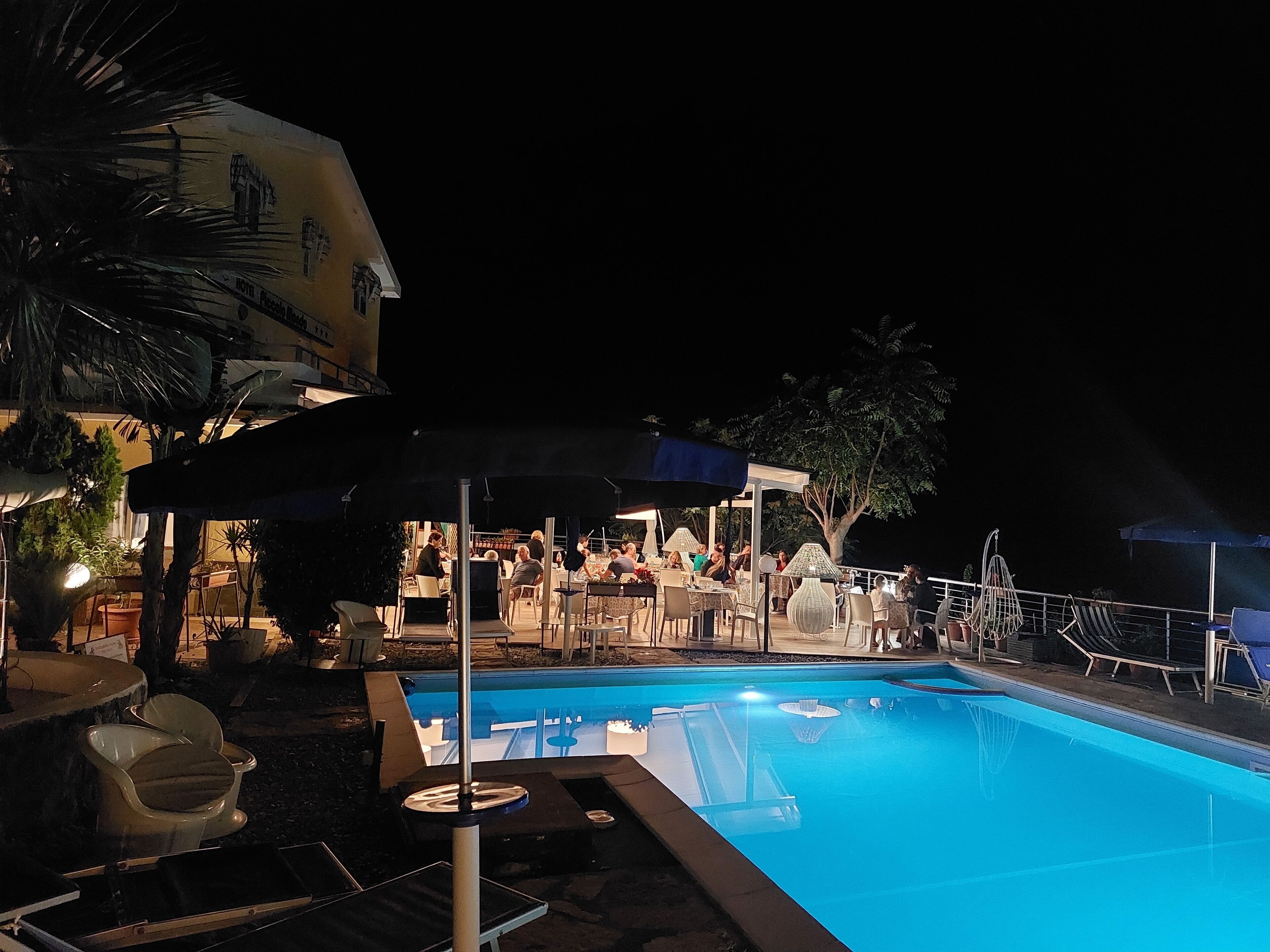 A nighttime pool area with lounge chairs and umbrellas, surrounded by palm trees and a building with a mural.