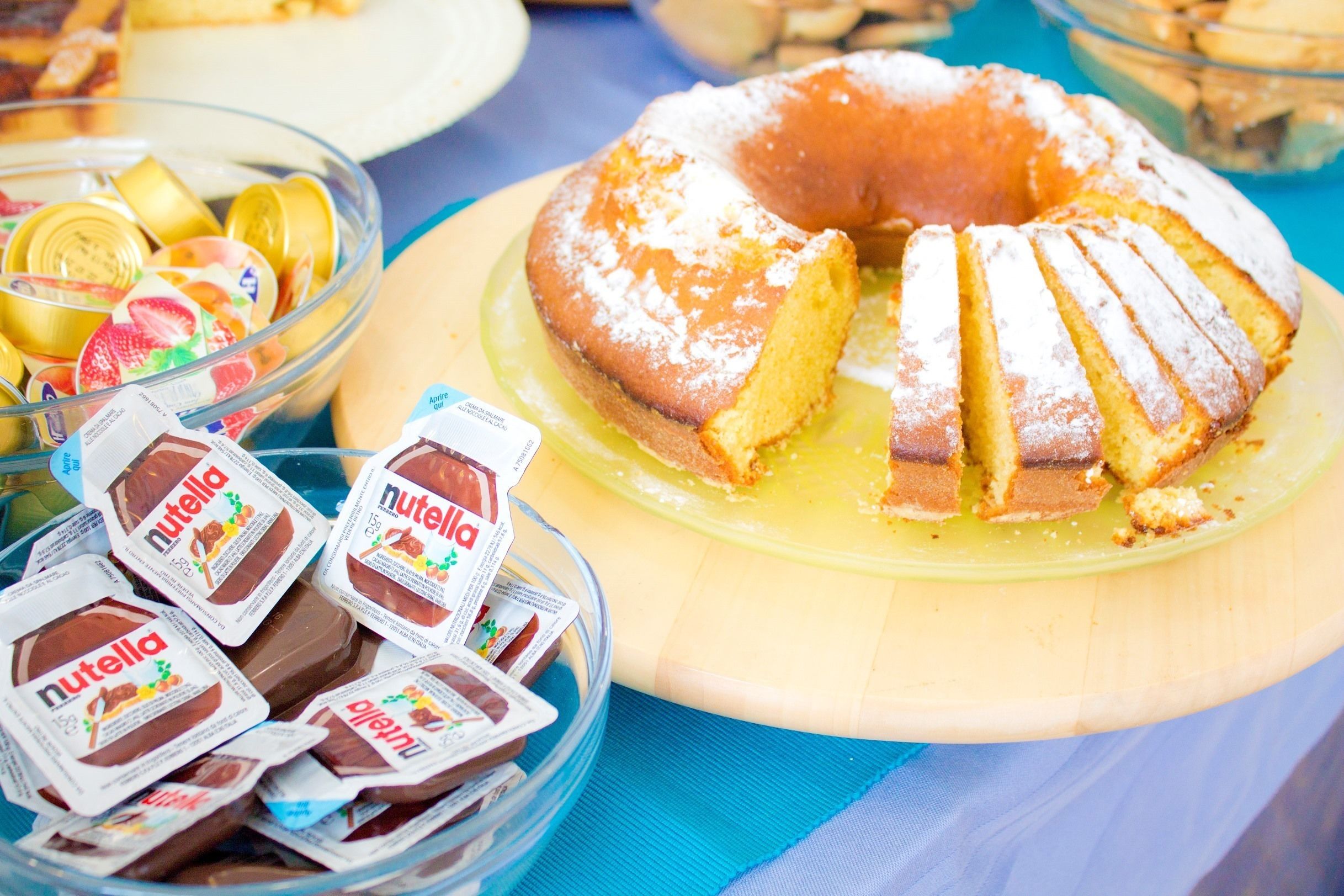A bundt cake with Nutella on a wooden board, surrounded by bowls of fruit and Nutella packets.