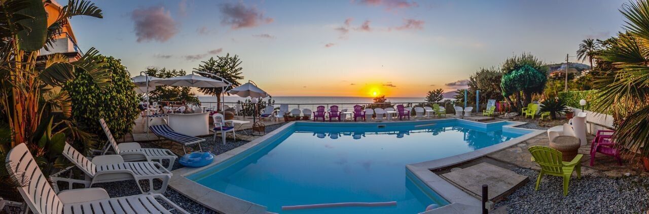 A pool area with lounge chairs and umbrellas, overlooking a sunset over the ocean.