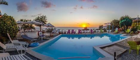 A pool area with lounge chairs and umbrellas, overlooking a sunset over the ocean.