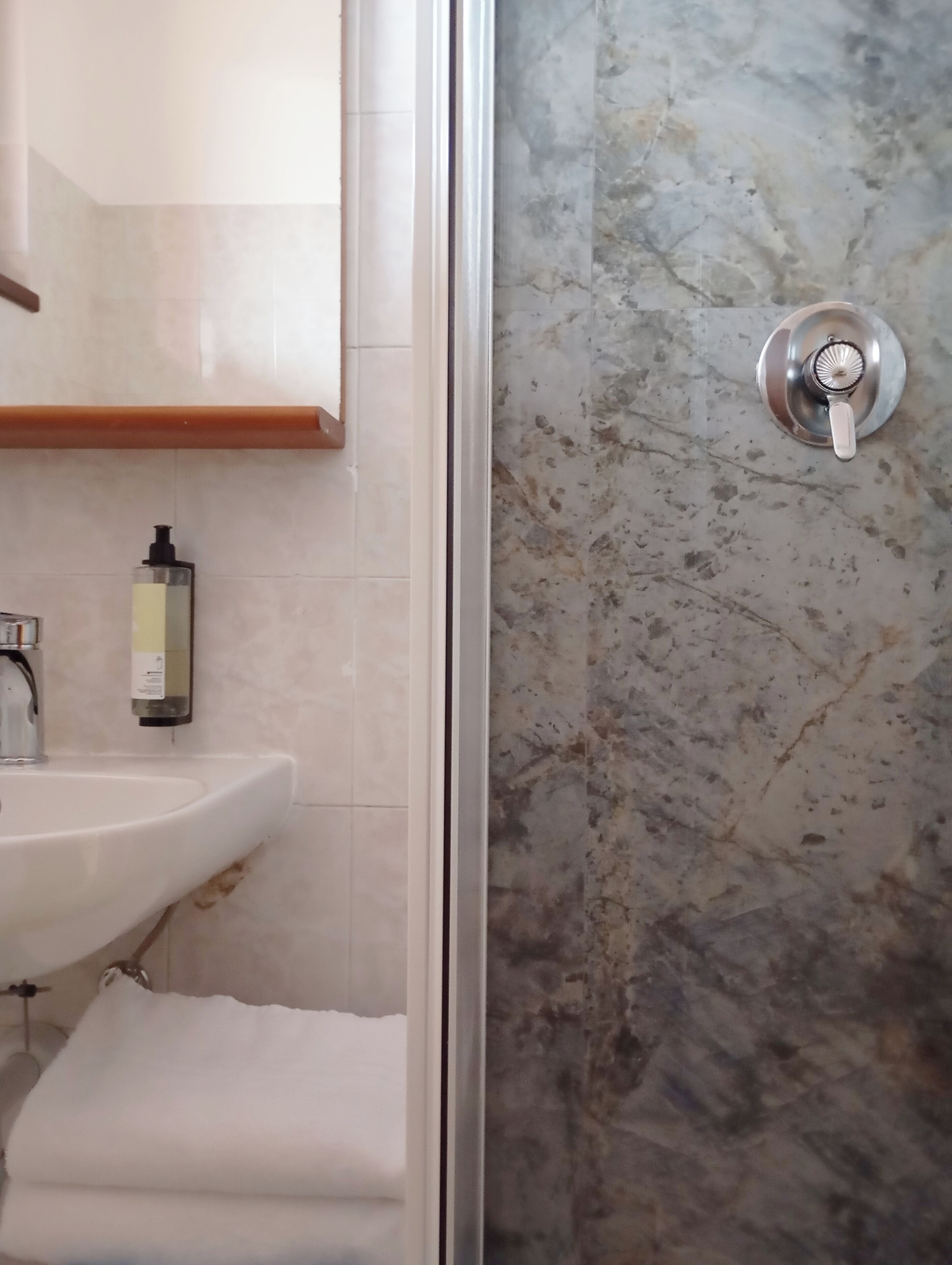 Une salle de bain avec un lavabo blanc, un miroir, un distributeur de savon et une douche avec un mur en marbre.