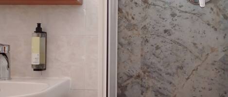 Une salle de bain avec un lavabo blanc, un miroir, un distributeur de savon et une douche avec un mur en marbre.