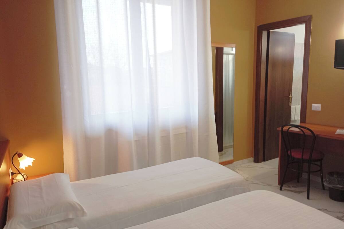 A hotel room with two single beds, a desk with a chair, a lamp, and a door.