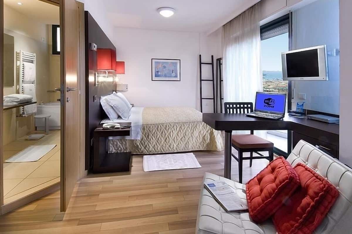 A hotel room with a bed, a desk with a laptop, a TV, and a view of the sea.