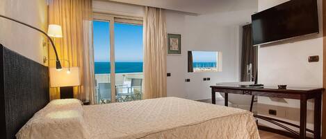 Habitación de hotel con cama, escritorio, televisión, teléfono y ventana con vista al mar.