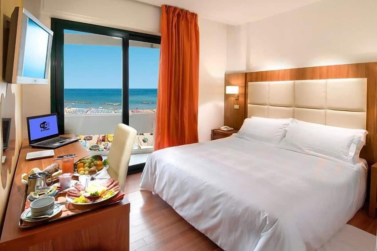 A hotel room with a bed, a desk with a laptop, a tray of breakfast, and a view of the beach.