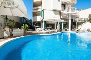 Una piscina con sillones de descanso y sombrillas, rodeada por un edificio con balcones y una escalera.